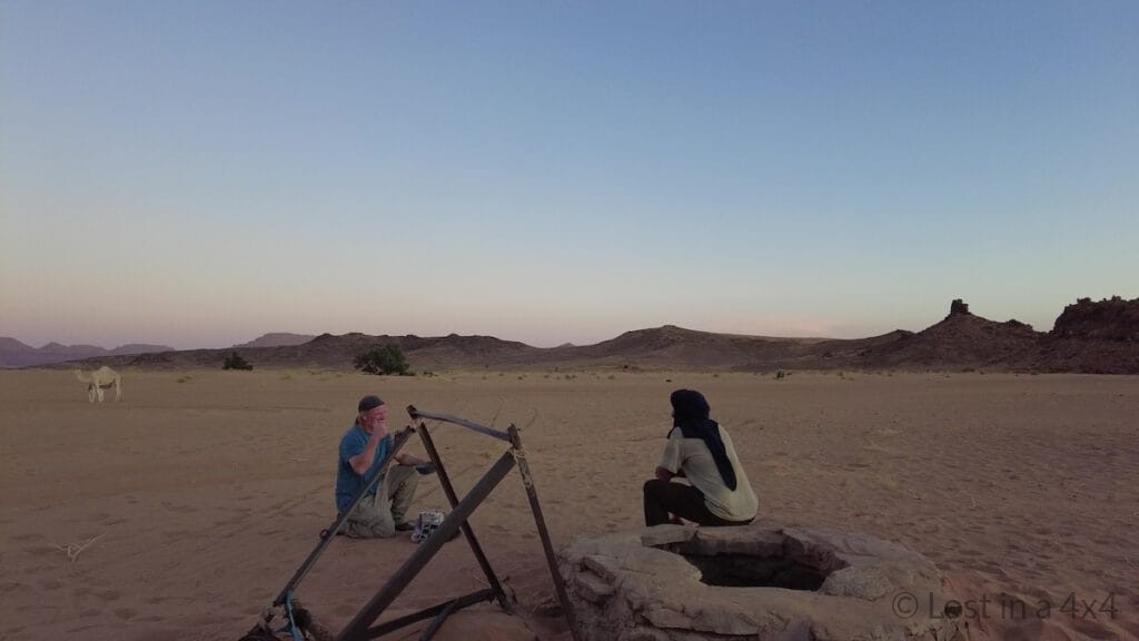 Brett and the camel herder standing next to the desert well, happy after the baby camel rescue, while the baby camel nurses from its mother in the background in Morocco
