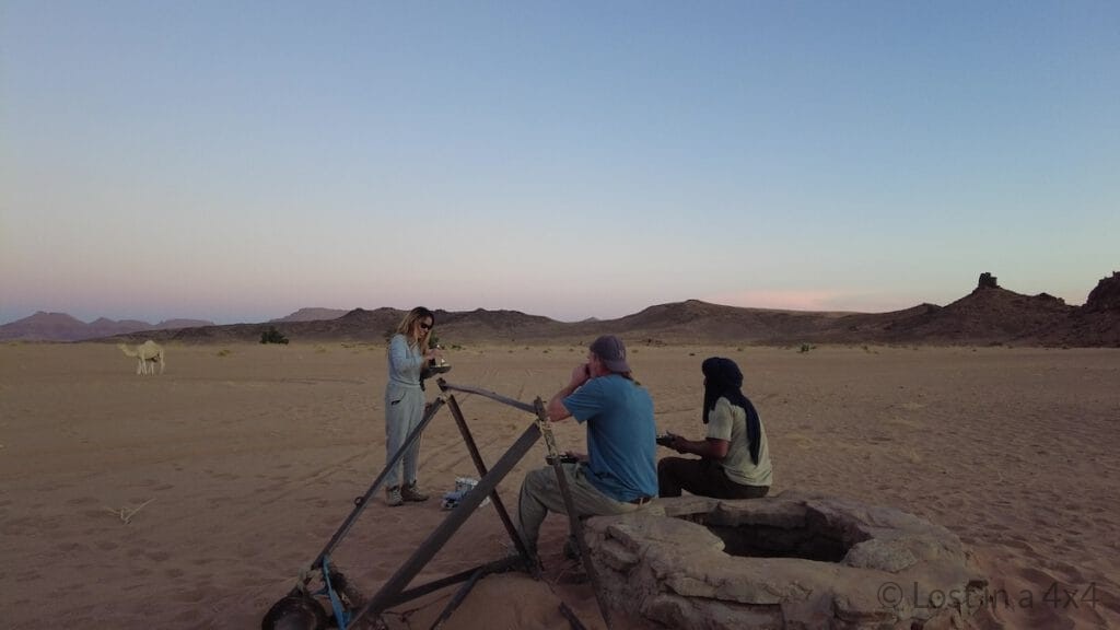 Me, Brett, and the camel herder sitting by the desert well having dinner after rescuing a baby camel, while the baby camel nurses from its mother in the background in Morocco