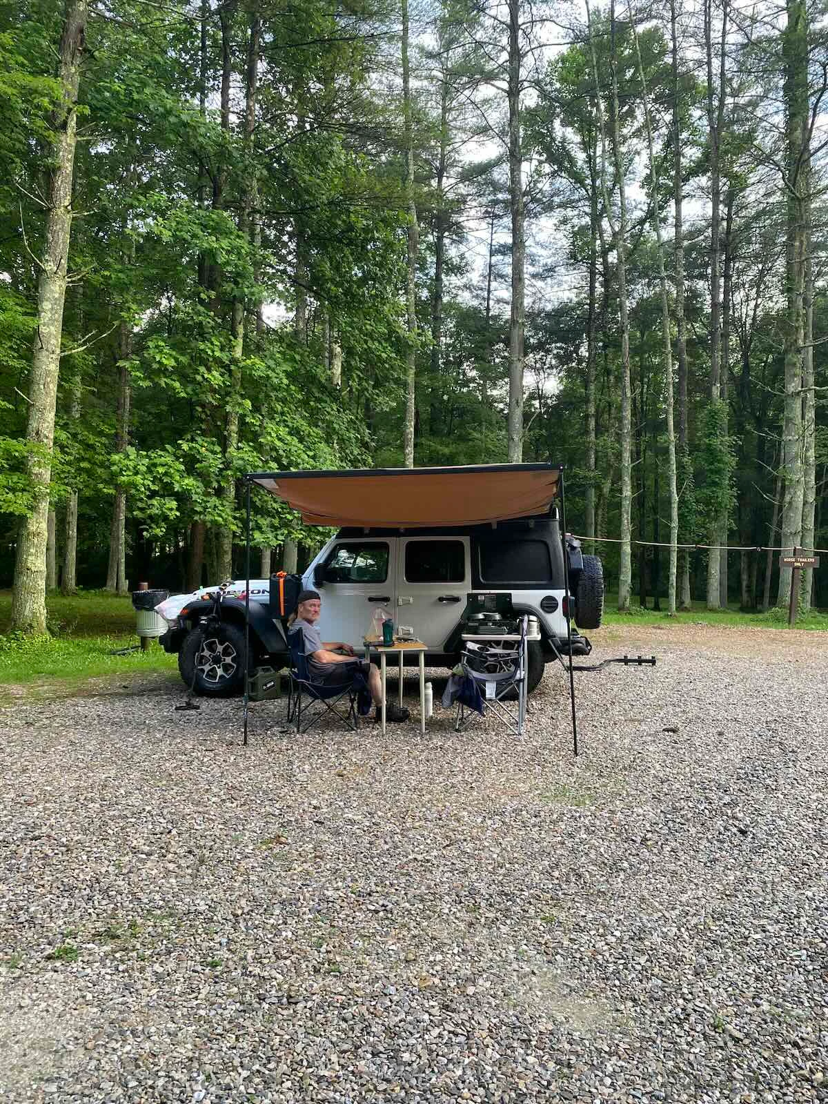 Overland camping setup with an ARB Touring Awning, DIY camp table, and camping chairs beside a Jeep Wrangler Rubicon.