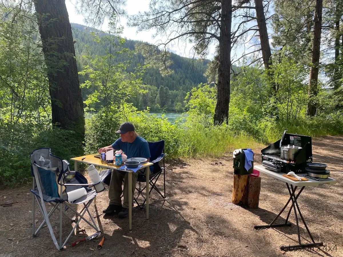 Brett sitting at DIY camping table with Coleman stove, chairs, and water jug at river wild camp in Montana