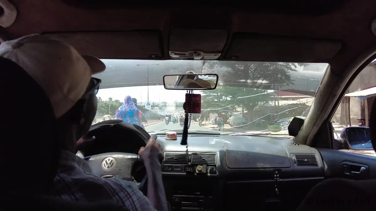 View from inside a taxi in Conakry, Guinea showing the driver, front seats, windshield, and the road ahead