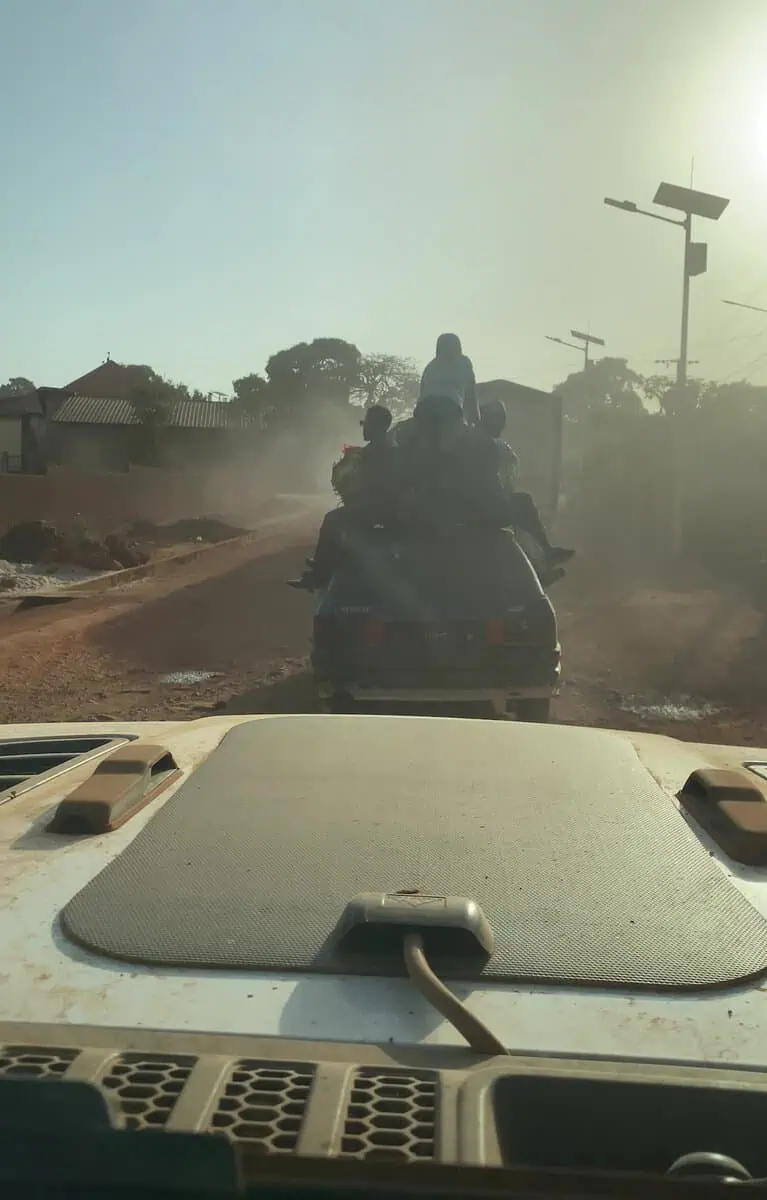 Car carrying passengers sitting and standing on roof while driving dirt road in Guinea