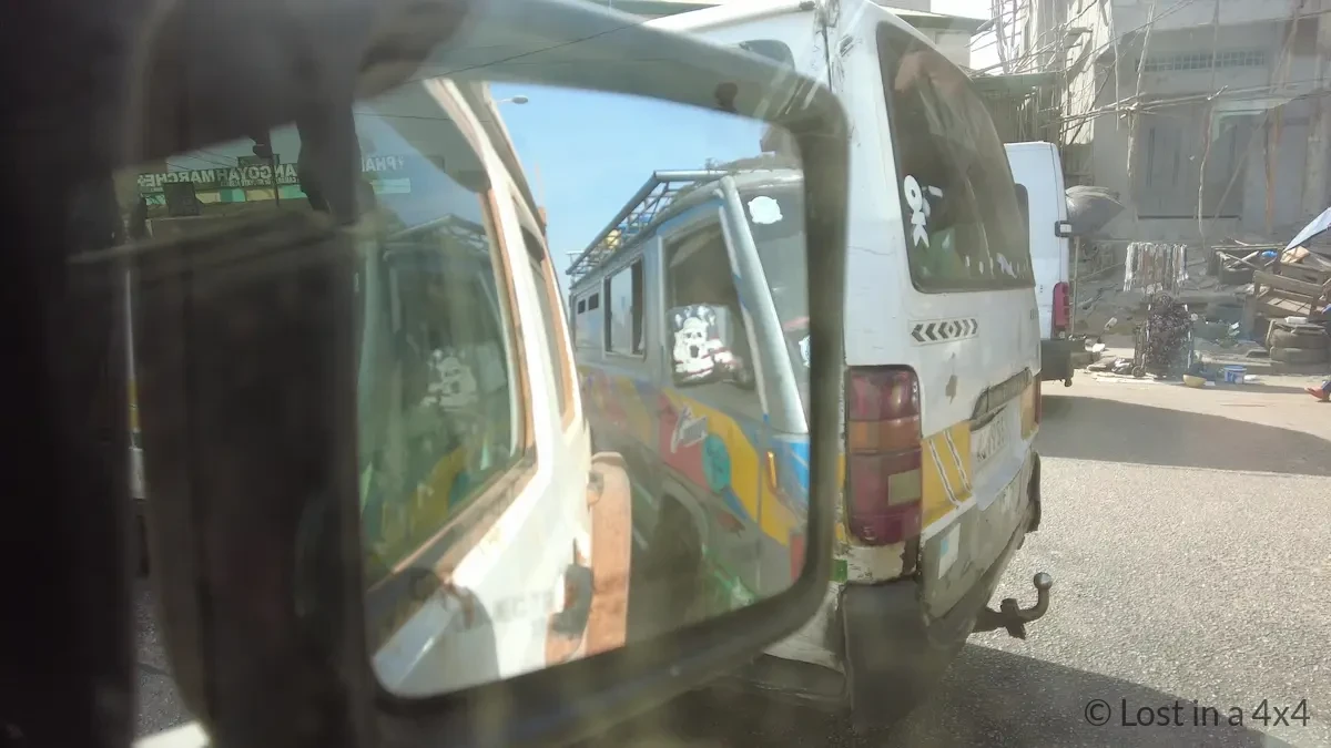 View from Jeep side mirror showing a minibus driving extremely close behind in Conakry, Guinea