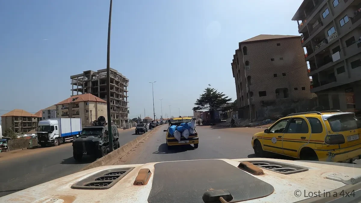 View from inside a Jeep Wrangler windshield showing a military convoy driving on the opposite side of the road in Conakry, Guinea