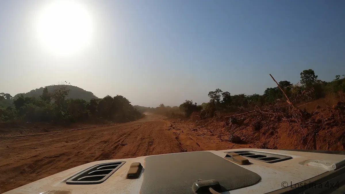 Inside windshield view from our Jeep Wrangler Rubicon showing the road ahead to Conakry, Guinea, where deteriorating tarmac transitions into dirt sections with visible potholes along the overland route through West Africa