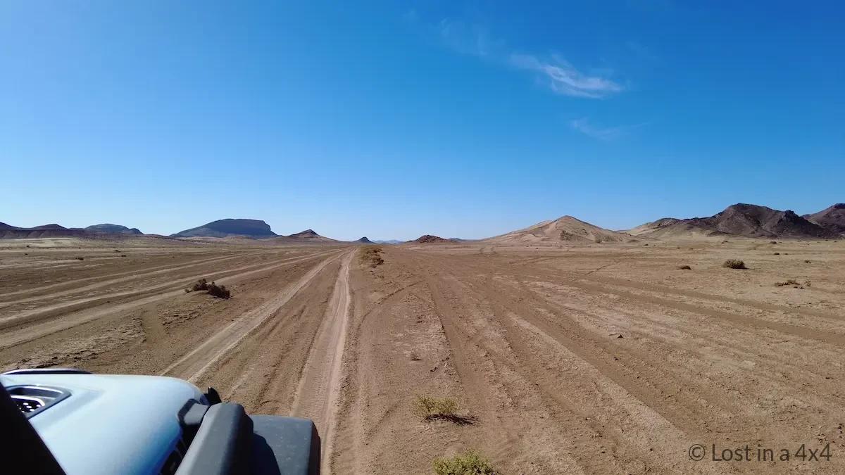 This photo shows the view from inside a 4x4 Jeep driving the remote desert road between Rissani and Ramlia in Morocco. The track stretches across flat sandy terrain with dirt tracks cutting through the landscape, while mountains rise in the distance.