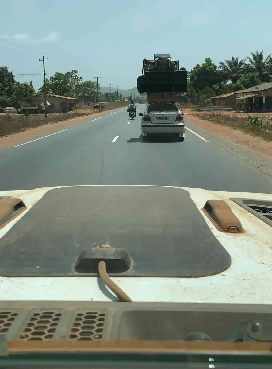 View from Jeep windshield of car carrying two couches and wood panels on roof on tarmac road to Conakry Guinea