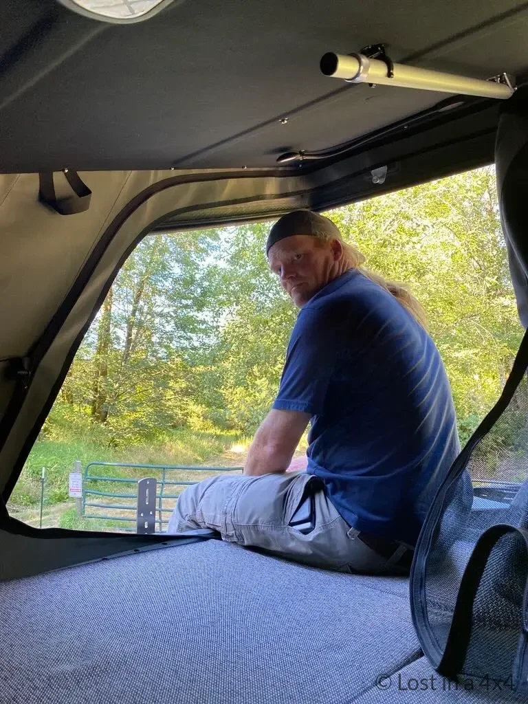 Brett sitting inside an Ursa Minor pop-top Jeep camper with legs hanging out of the open window, showing interior bed cushions.