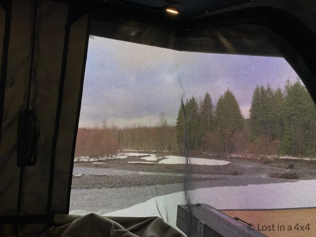 Interior view of the left-side window and mosquito net inside an Ursa Minor pop-top camper during snowy camping conditions.