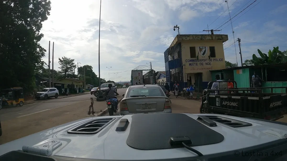 Dash cam view approaching the Noé border crossing from Ivory Coast with the Commissariat de Police Mixte de Noé building visible on the right side of the road.